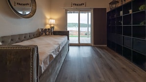 Un dormitorio con cama, espejo, lámpara, vistas al exterior y repisas con objetos decorativos.