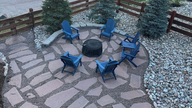 Un patio con baldosas de piedra, sillas Adirondack azules, un hogar para fuego y vista al lago.
