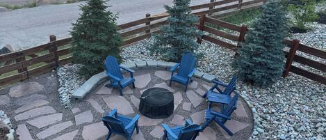 A patio with stone tiles, blue Adirondack chairs, a fire pit, and a view of a lake.