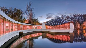 Una estructura arquitectónica moderna con un puente curvo sobre una masa de agua reflectante, rodeada de árboles.
