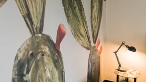 Una obra de arte con cactus en una pared, una lámpara de escritorio y una pequeña mesa con estampado floral.