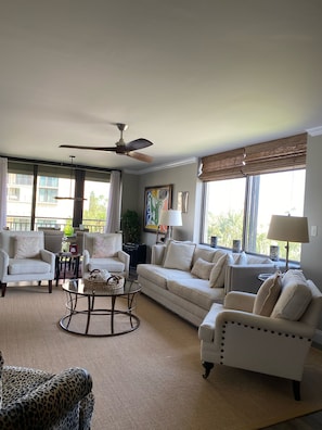 A spacious living room with a ceiling fan, large windows with blinds, a sofa set, a coffee table, and a leopard-print armchair.