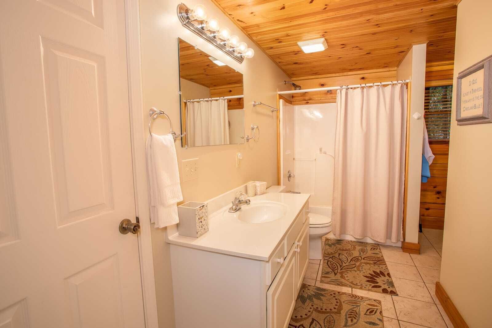 A bathroom with a wooden ceiling, a white sink, a mirror with lights, a shower curtain, and a patterned rug.