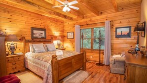 A cozy wooden cabin bedroom with a bed, nightstands, a lamp, a chair, and a window with curtains. - Spacious Boone log cabin with 4 bedrooms, 3 baths, river access and game room (Boone)