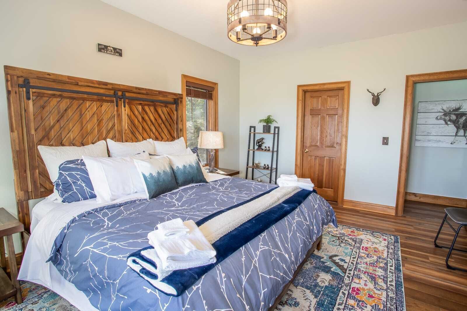 A bedroom with a large bed, wooden headboard, a chandelier, a door with a deer head, and a rug.