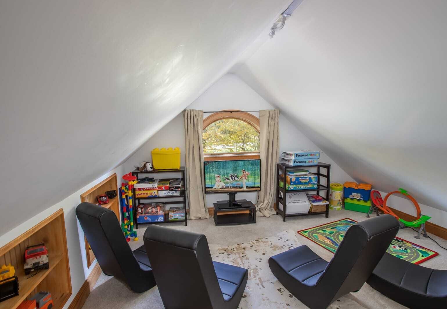 Ein gemütliches Zimmer im Dachgeschoss mit schräg stehender Decke, einem großen Fenster mit Vorhängen, einem Fernseher und verschiedenen Kinderspielen und -spielzeug.