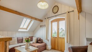 Une pièce confortable de style loft, avec un plafond en bois, une grande fenêtre avec des rideaux, une table en bois ornée d’un vase de fleurs et un canapé en cuir marron.