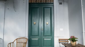 Une porte verte avec deux poignées rondes, une chaise en osier et un banc en bois sur lequel est posée une plante en pot.