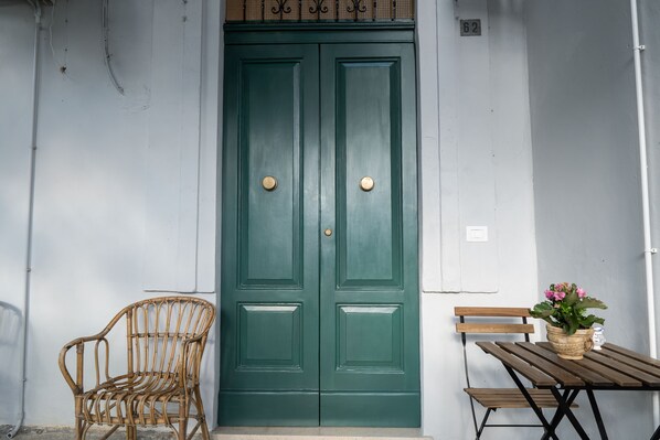 Una puerta verde con dos pomos redondos, una silla de mimbre y un banco de madera con una planta en maceta.