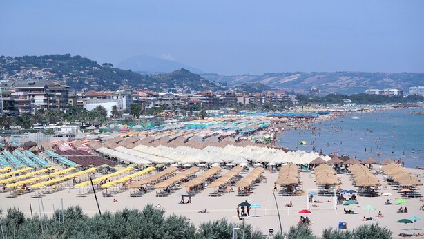 Une plage bordée de rangées de transats et de parasols, un ciel dégagé et des bâtiments en arrière-plan.