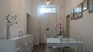 Une cuisine moderne avec des meubles blancs, une table à manger au plateau en verre et des chaises blanches. On peut voir un vase avec des branches séchées et une orchidée rose sur le comptoir.