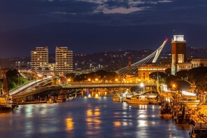 Vista nocturna de una ciudad con un puente atirantado destacado, un río con barcos y edificios iluminados al fondo.