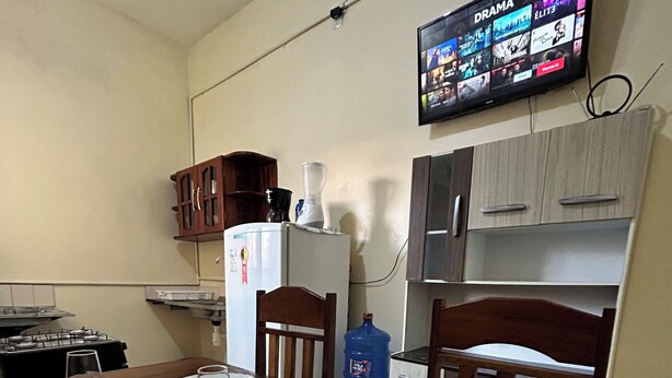 Un comedor con una mesa dispuesta para cuatro personas, un refrigerador, un dispensador de agua y un televisor montado en la pared mostrando un canal de drama.