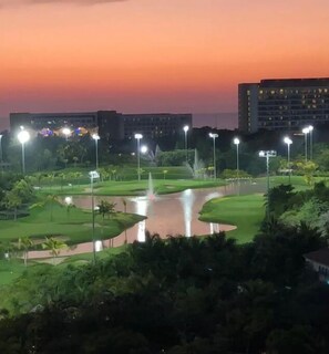 Een golfbaan in de schemering met een waterpartij, verlicht door lantaarns, omringd door gebouwen.