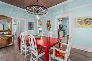 Une salle à manger avec une table rouge, des chaises blanches, un lustre et une cuisine en arrière-plan.