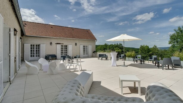 A spacious outdoor patio with modern white furniture, including sofas and chairs, and a large umbrella providing shade.