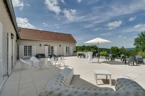 Un amplio patio al aire libre con mobiliario blanco moderno, incluyendo sofás y sillones, y una gran sombrilla que proporciona sombra.