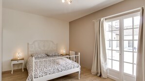 A bedroom with a white metal bed frame, a bedside table with a lamp, and a view through a large window with curtains.