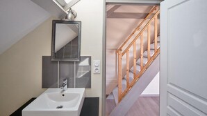 A bathroom with a white sink, a mirror, and a light fixture above it. There is a wooden staircase leading to another room.