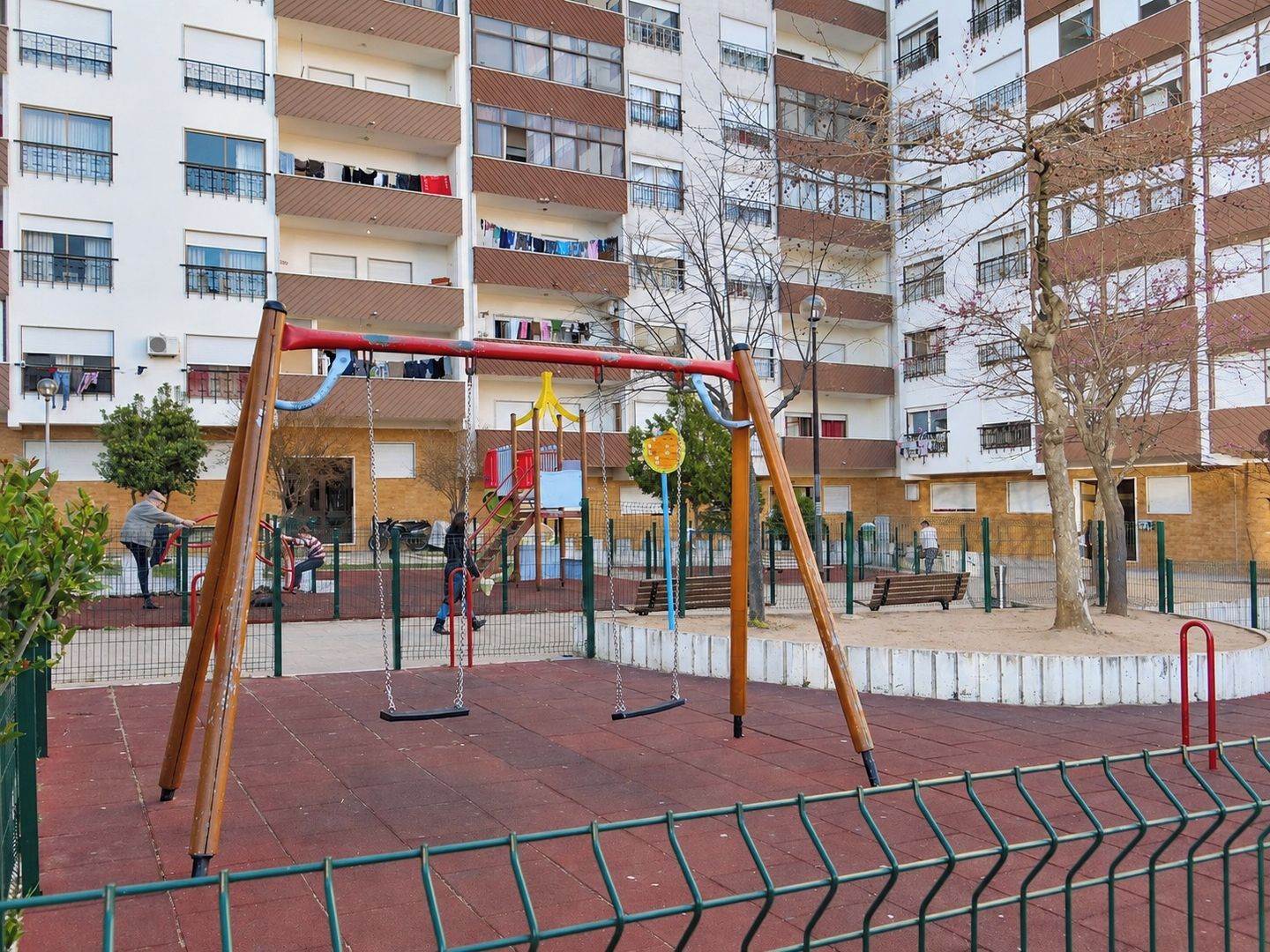Une aire de jeux avec des balançoires et une structure d’escalade, entourée d’une zone délimitée par une clôture, comprenant des bancs et un bâtiment en arrière-plan.