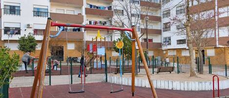 Un patio de juegos con columpios y una estructura para escalar, rodeado por una zona vallada con bancos y un edificio al fondo.