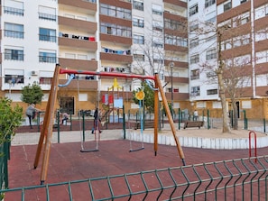 Ein Spielplatz mit Schaukeln und einer Kletterkonstruktion, umgeben von einem eingezäunten Bereich mit Bänken und einem Gebäude im Hintergrund.