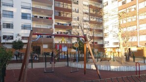 Ein Spielplatz mit Schaukeln und einer Kletterkonstruktion, umgeben von einem eingezäunten Bereich, mit einem Gebäude im Hintergrund.