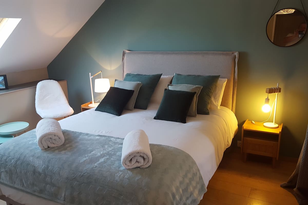 Uma cama bem arrumada com duas toalhas enroladas, uma mesa de cabeceira com abajur e um espelho fixado na parede.
