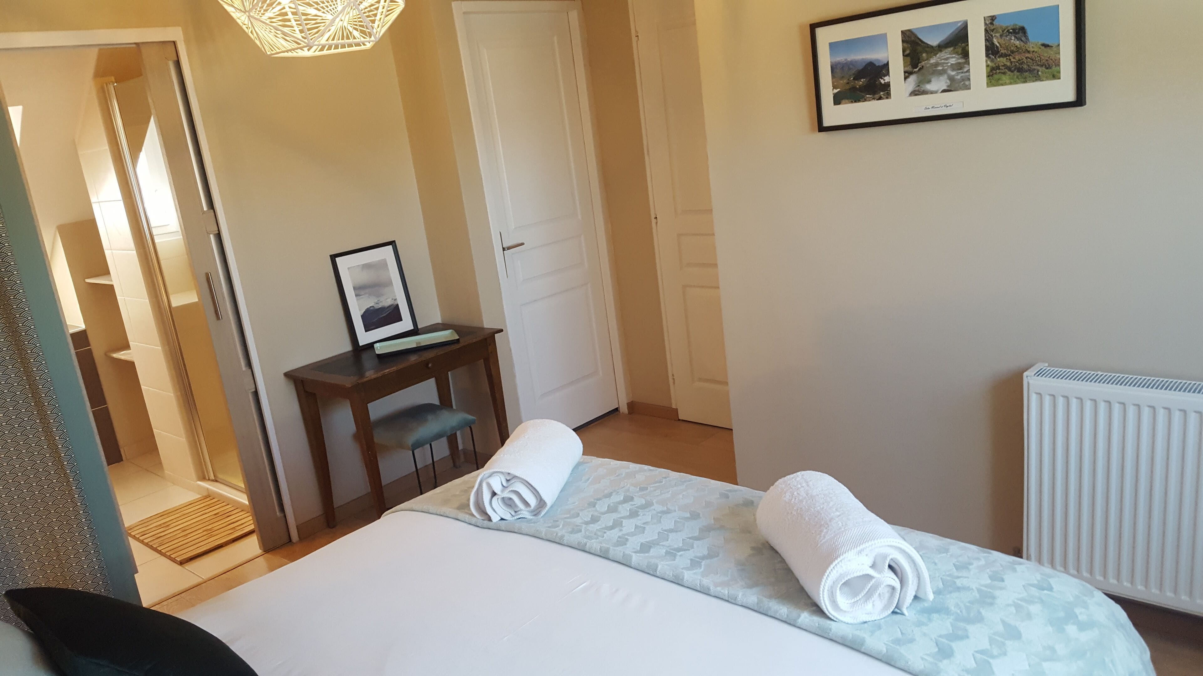 A hotel room with a bed, a desk with a computer, a framed picture on the wall, and a radiator.