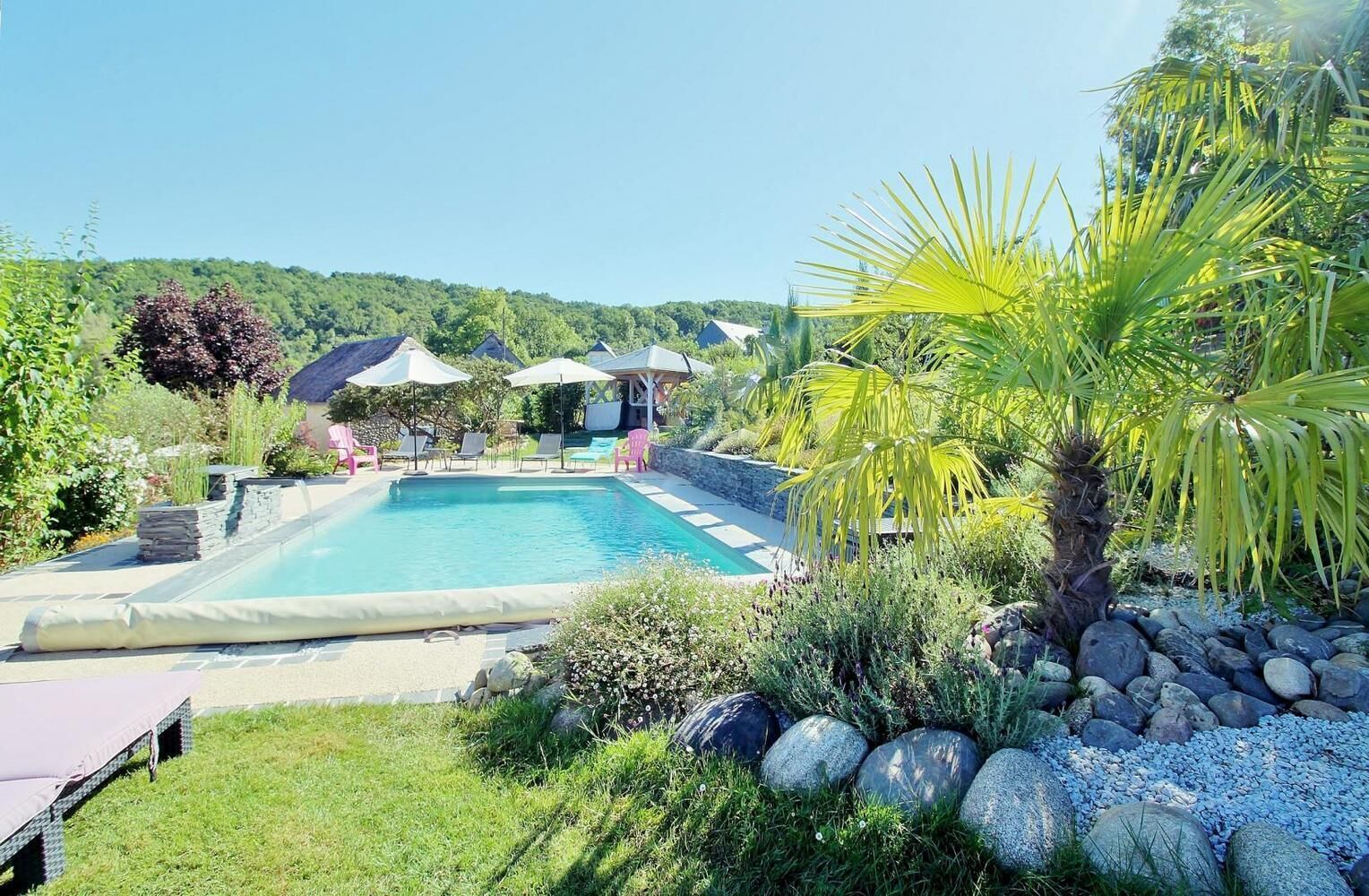 A rectangular pool with a stone border, surrounded by a grassy area with rocks and plants. There are lounge chairs and umbrellas in the background.