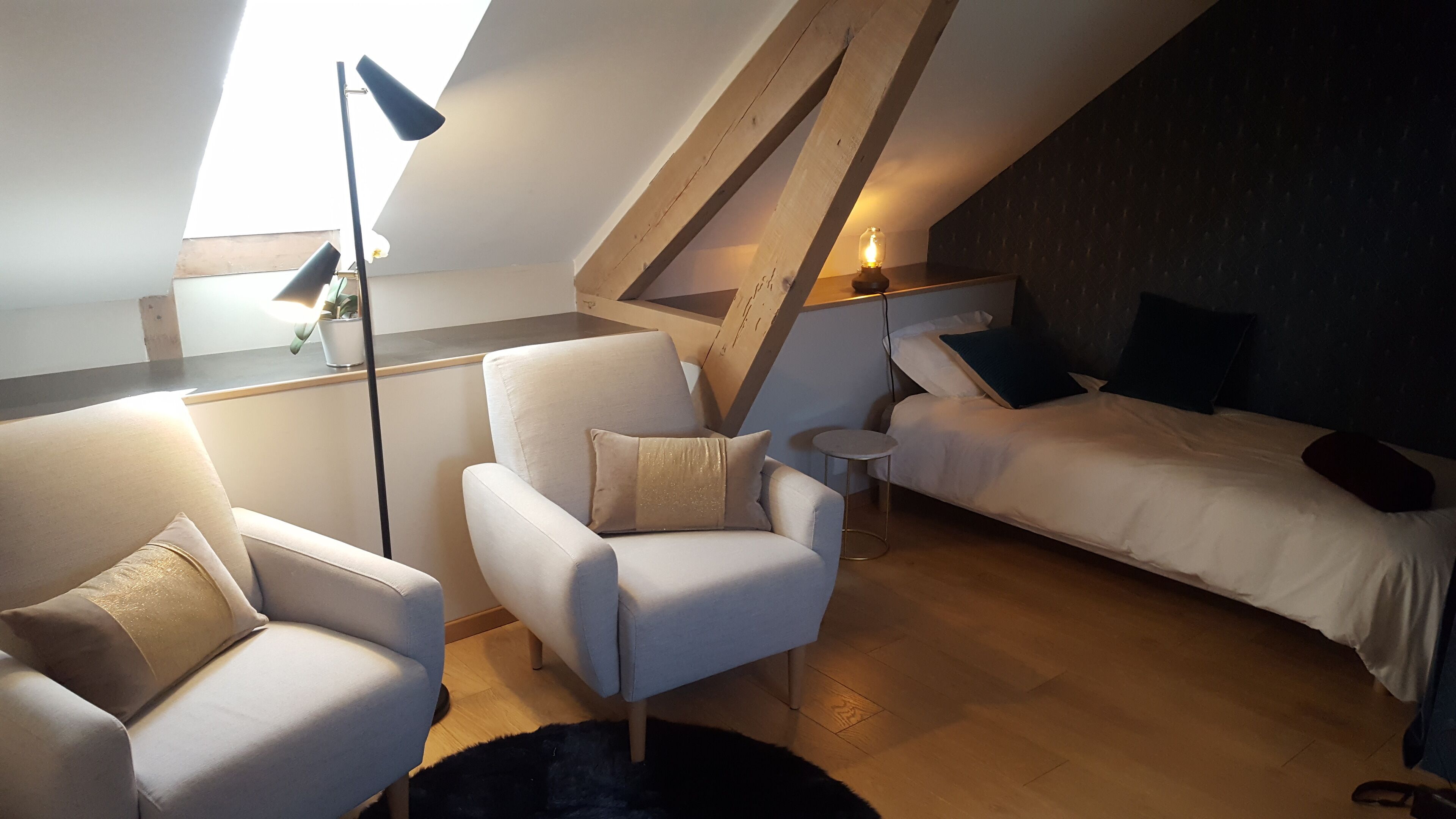 A cozy loft bedroom with a sloped ceiling, a bed, two armchairs, a small round table, and a floor lamp.