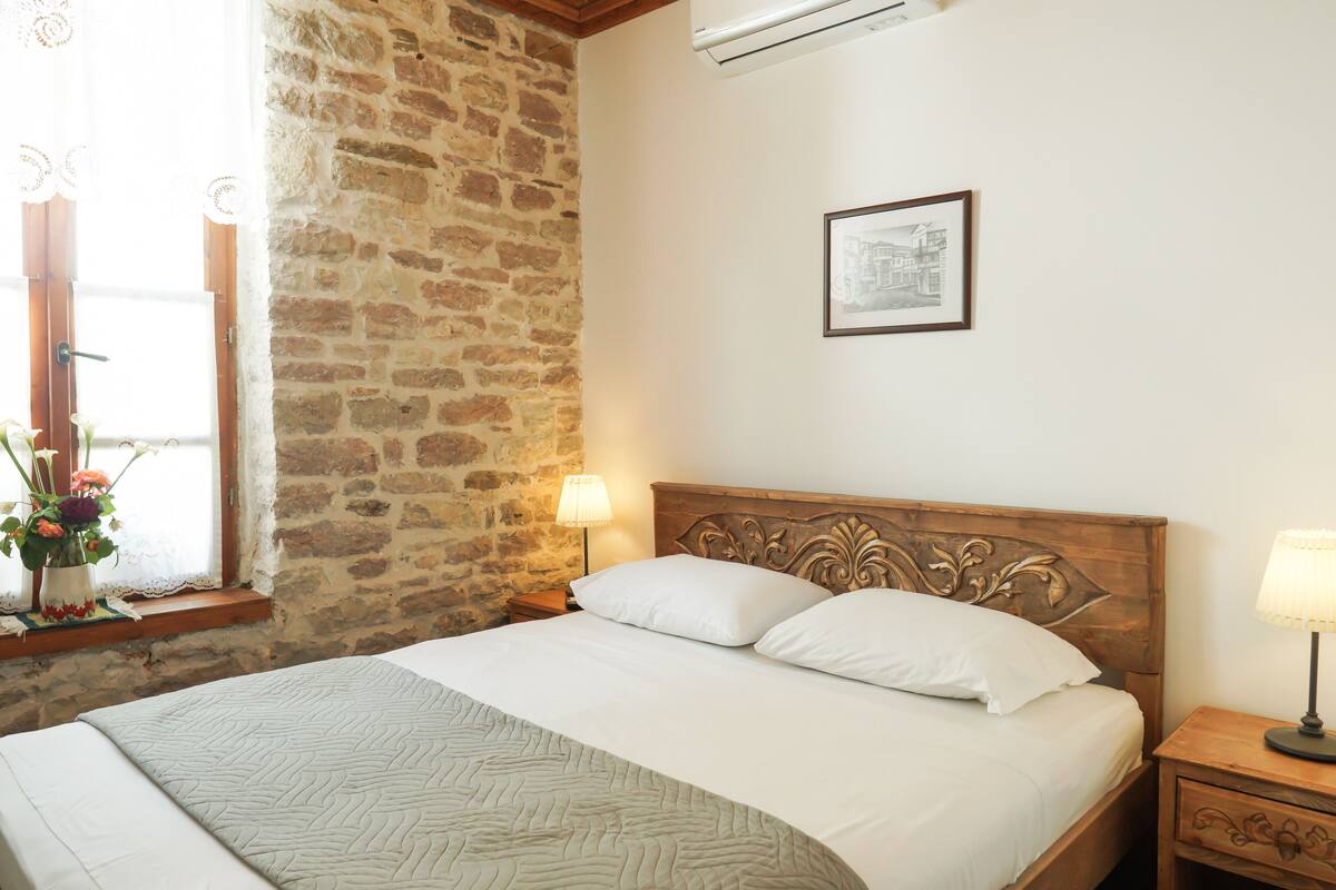 A bedroom with a stone wall, a bed with white linens, a nightstand with a lamp, and a framed picture on the wall.