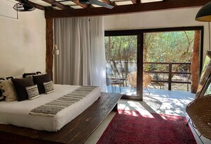 Un dormitorio con cama, una alfombra roja, un balcón con mobiliario de mimbre y vistas a árboles.