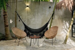 Un sillón hamaca colocado entre dos sillas de mimbre con una mesita redonda en un espacio exterior sombreado.