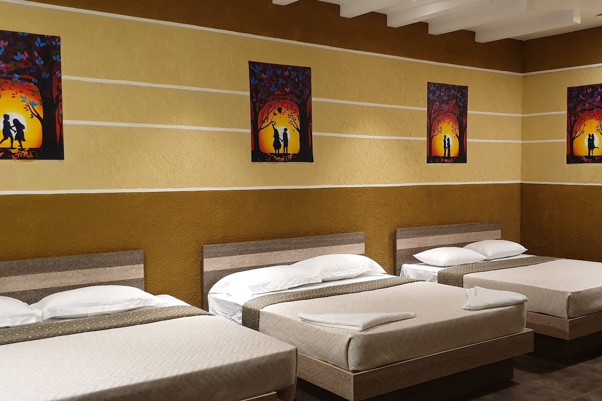A hotel room with four beds arranged in a row, each with white bedding and a wooden headboard. The walls are painted in a warm yellow tone, and there are three paintings of a couple under a tree with a sunset in the background.