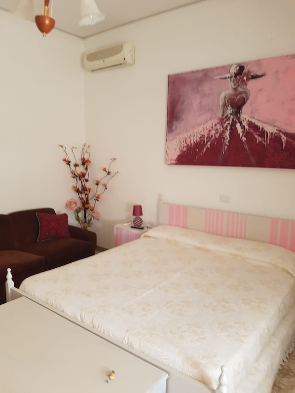 A bedroom with a large bed, a bedside table with a lamp, a small table, a brown sofa, and a wall-mounted air conditioner.