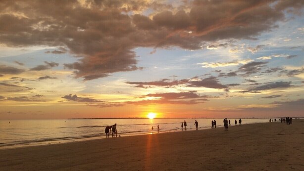 A beach scene with the sun setting over the horizon, people walking along the shore, and a distant pier. - Water Views, Pool, Private boat dock, Walking distance to Times Square (Fort Myers Beach)