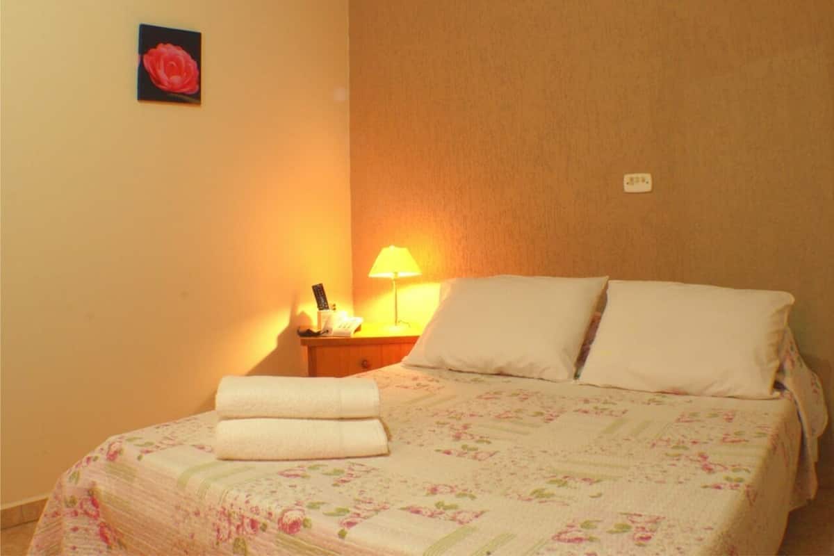Un lit bien fait, avec une literie à motifs floraux, une lampe de chevet et une seule rose rouge en décoration sur le mur.