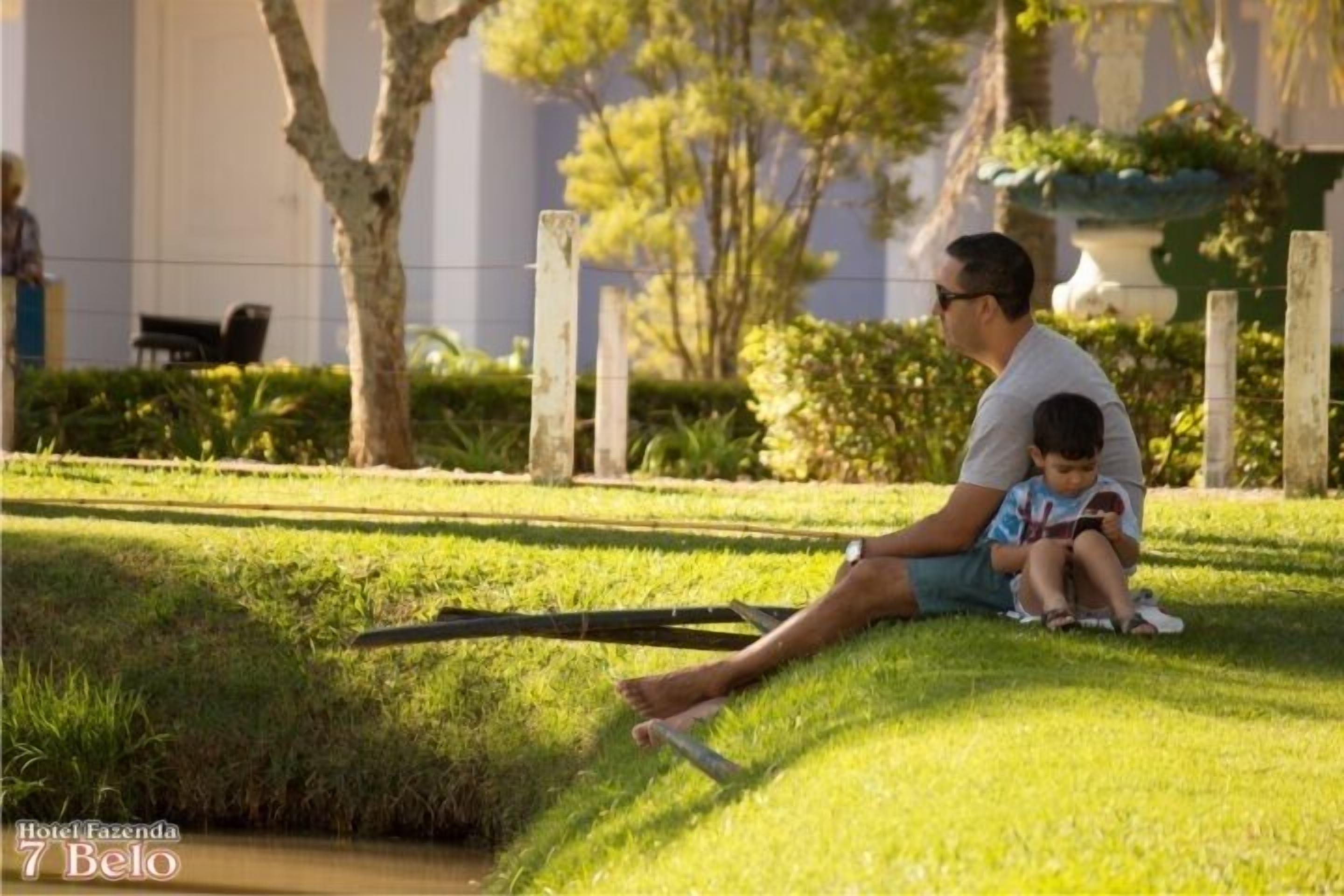 A man and a child sitting on grass in a park-like setting with a building in the background.