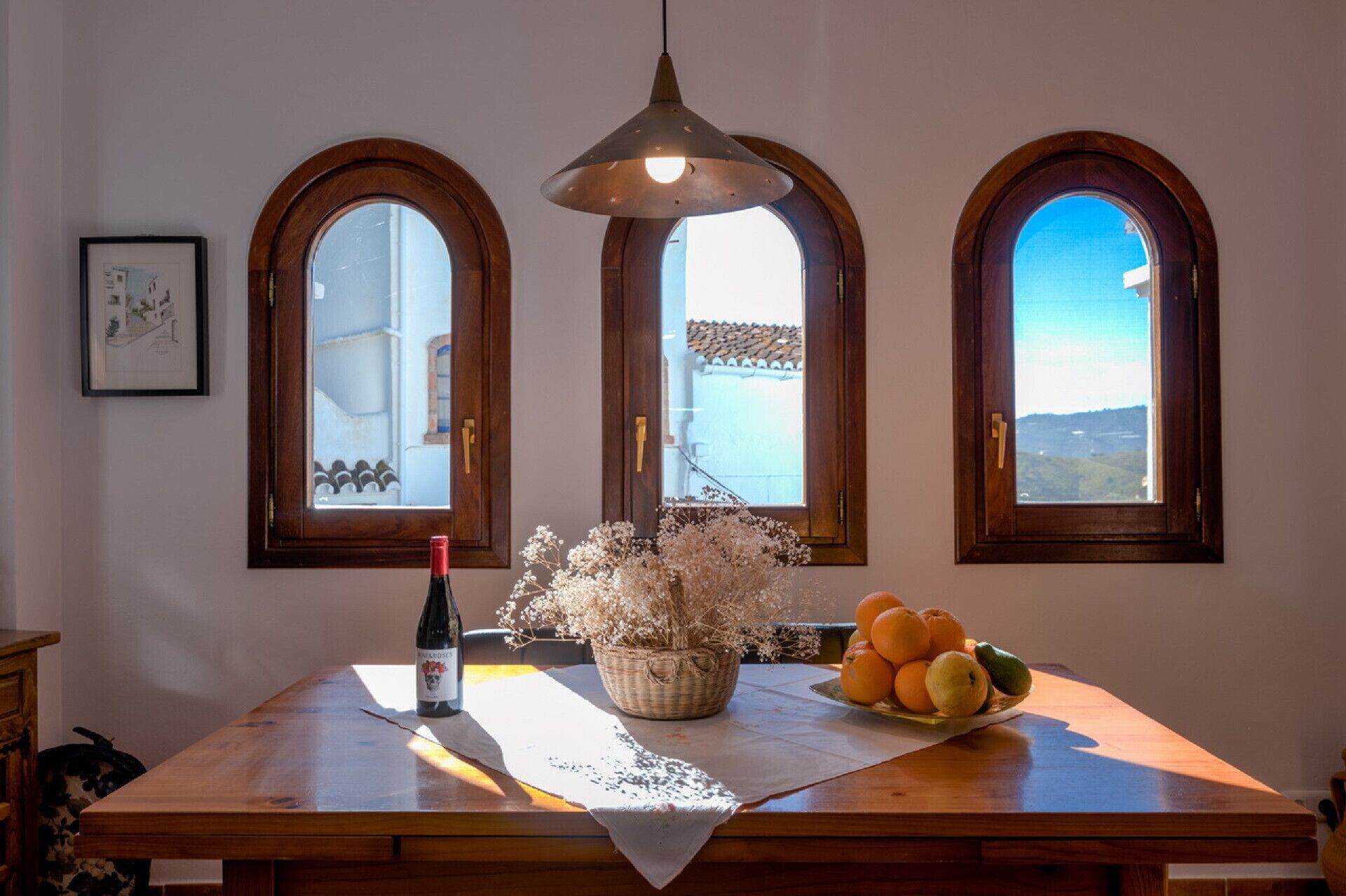 Ein Holztisch mit einer Weinflasche, einem Korb getrockneter Blumen und einem Obstteller. Drei Bogenfenster bieten Blick auf ein Gebäude und die Landschaft.