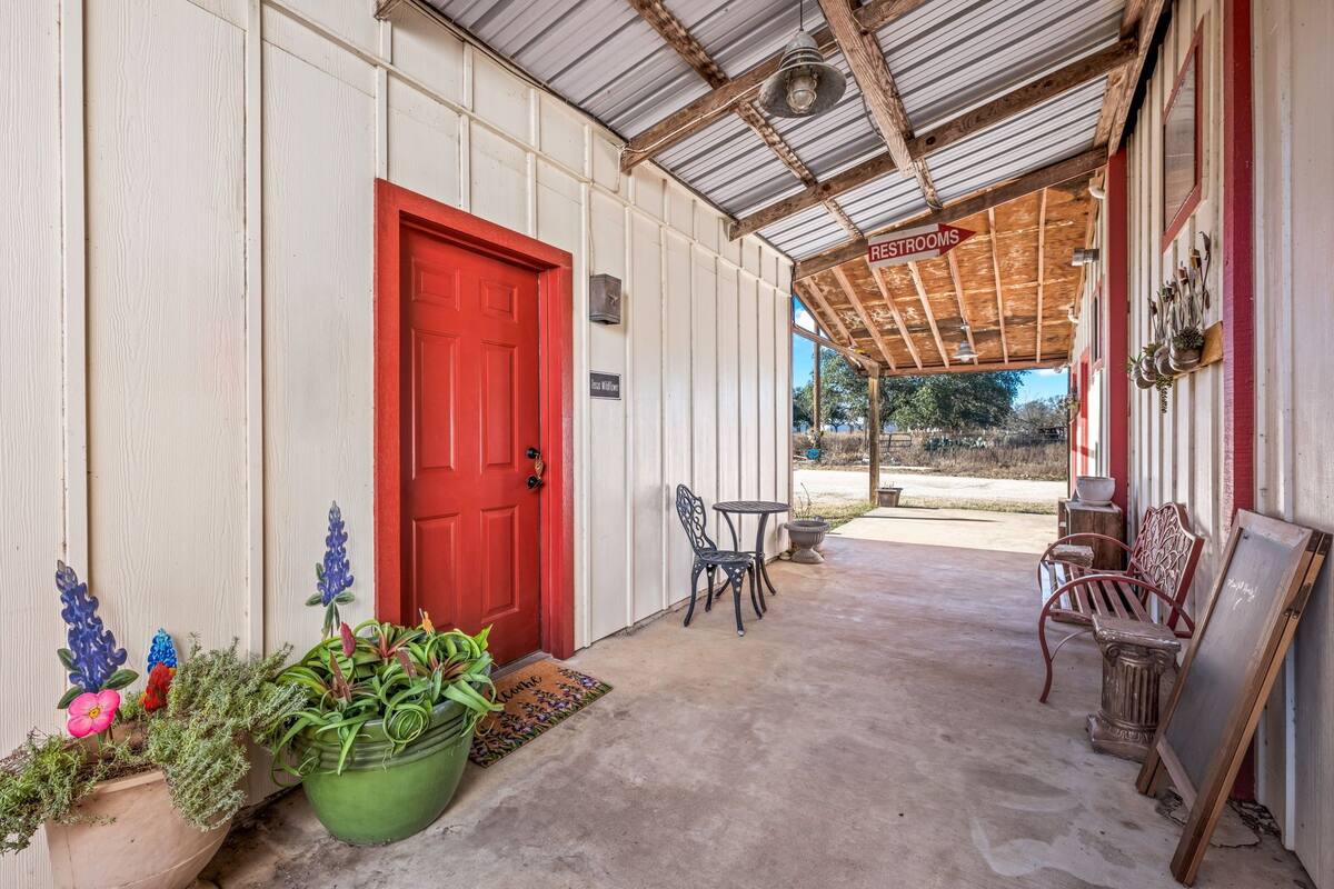 A covered porch with a red door, a sign indicating restrooms, a table with chairs, and potted plants.