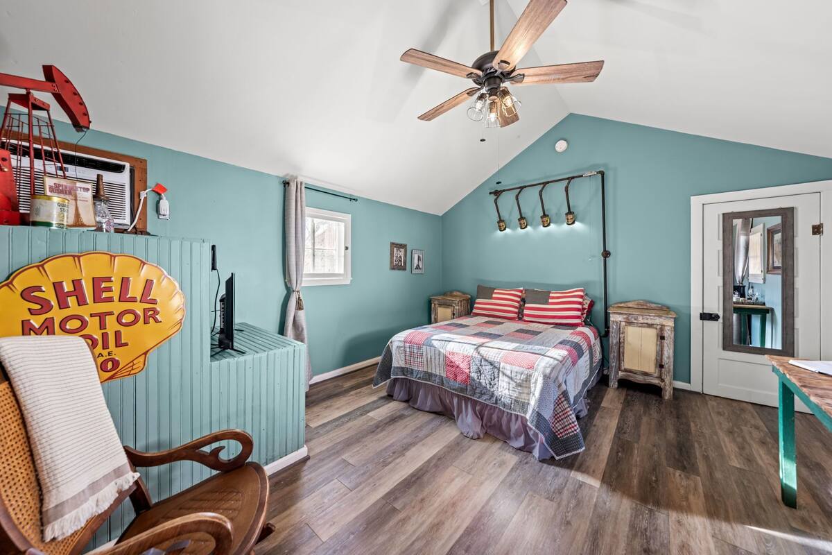 A bedroom with a bed, a ceiling fan, a wooden chair, a small table, and a wall-mounted shelf with various items.