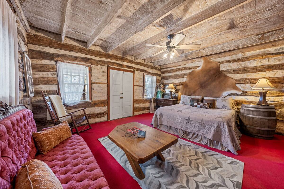A log cabin bedroom with a bed, a red carpet, a wooden coffee table, a leather sofa, and a ceiling fan.