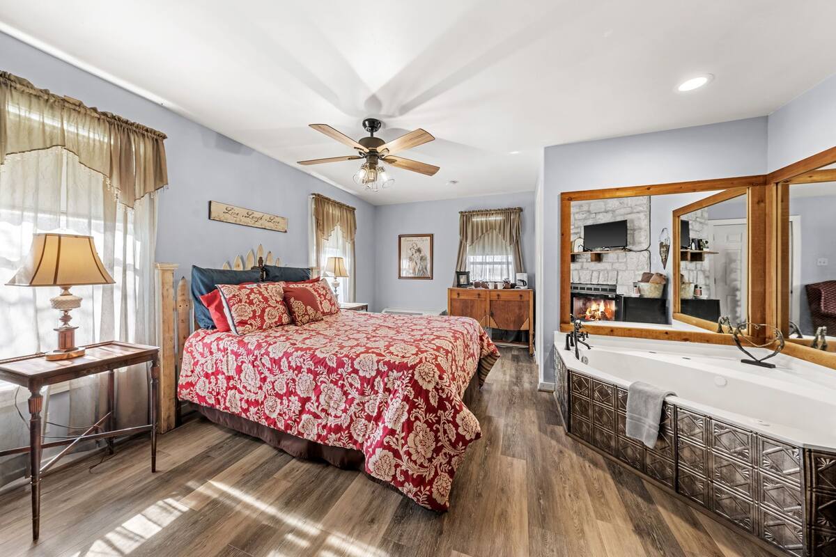 A bedroom with a large bed, a ceiling fan, a wooden dresser, a large mirror, and a bathtub.