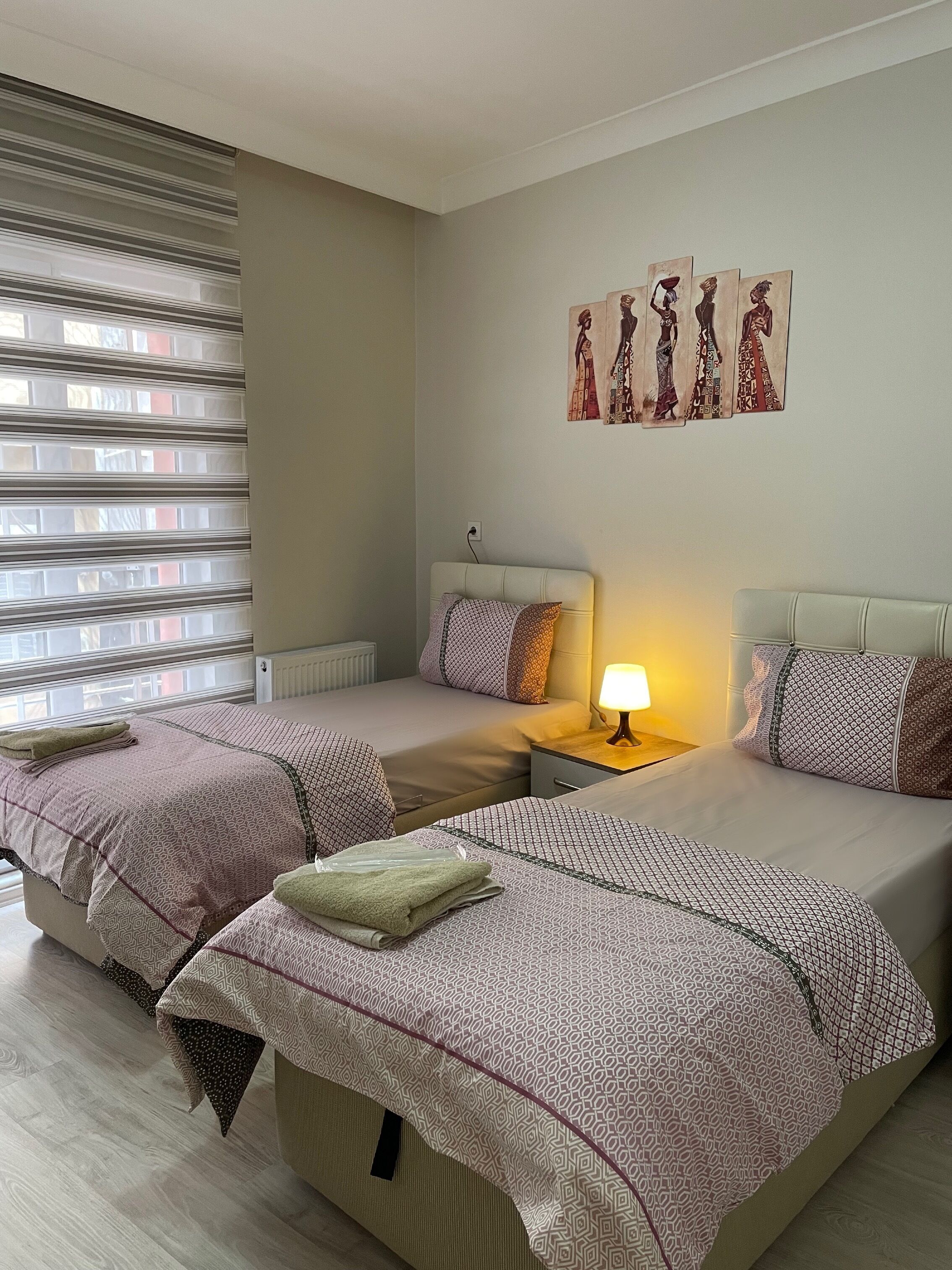 A hotel room with two beds, a window with blinds, a nightstand with a lamp, and artwork on the wall.