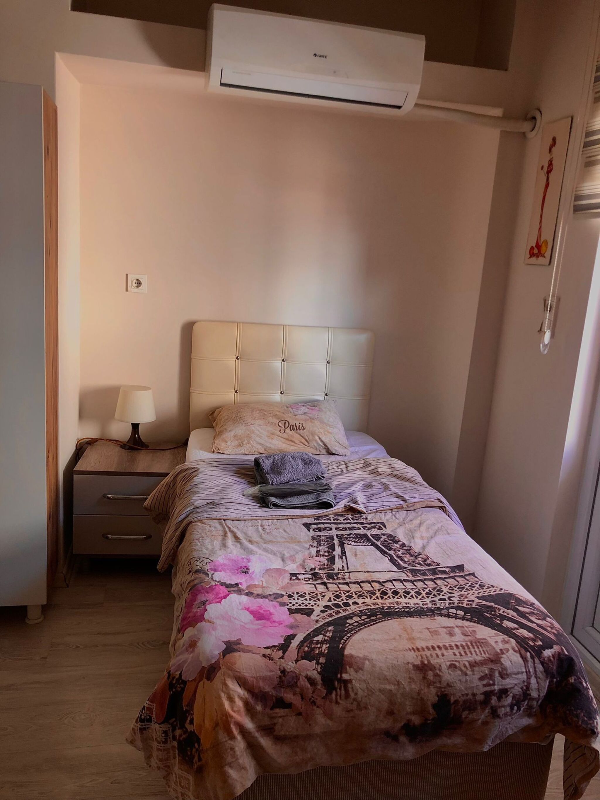 A bedroom with a bed, a nightstand, a lamp, an air conditioner, and a wall-mounted light fixture.
