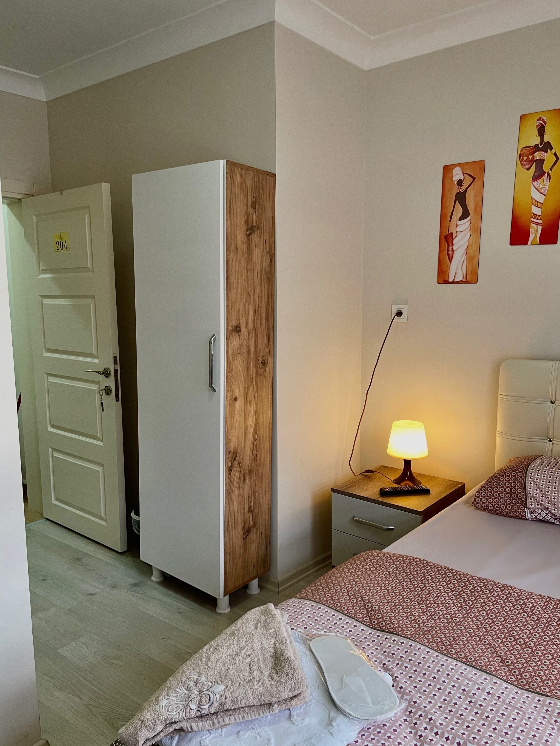 Habitación de hotel con cama, armario de madera, mesita de noche con lámpara y una obra de arte en la pared.