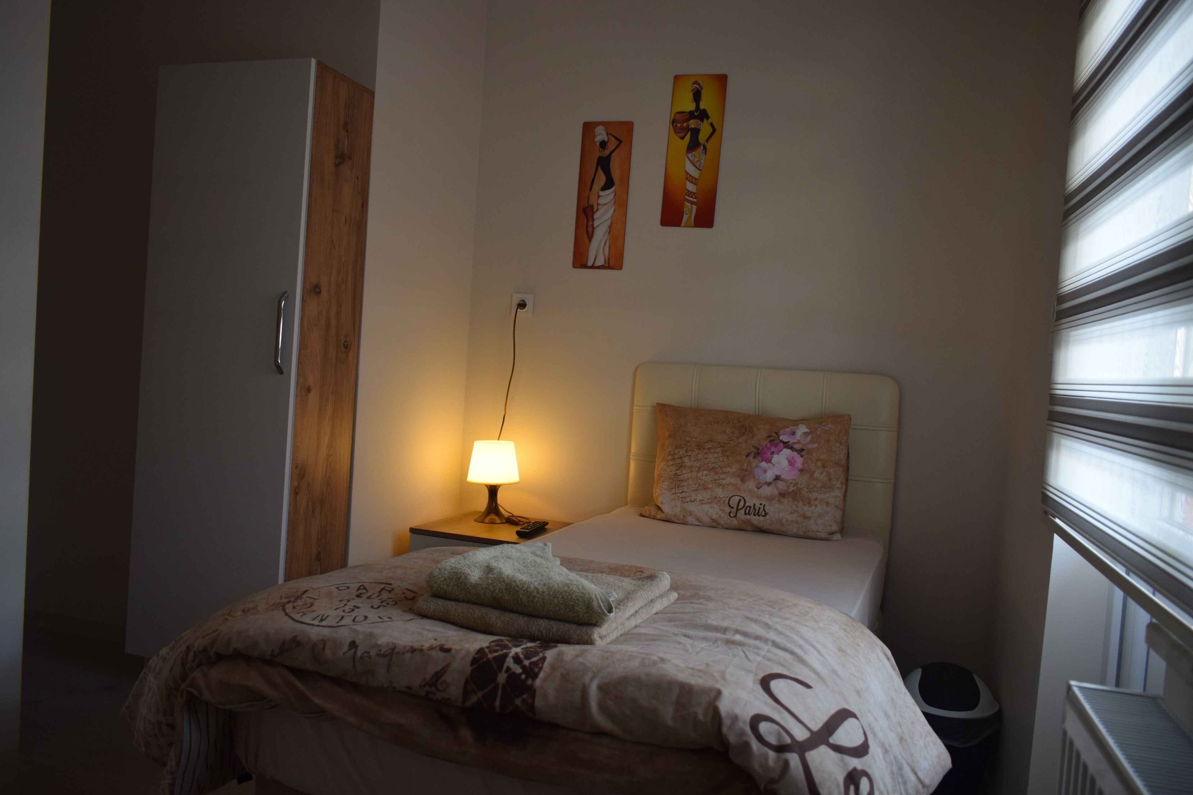 A bedroom with a bed, a nightstand with a lamp, a wooden wardrobe, and two wall art pieces.