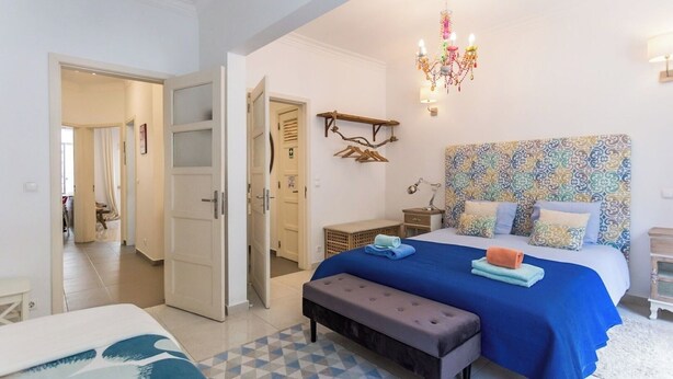 A bedroom with a large bed, a bench, a chandelier, and a patterned headboard. - 1-Bedroom Flat with Terrace, near Metro and Arena (Loures)