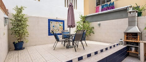 A patio with a table and chairs, a potted plant, a grill, and a tiled wall with a colorful tile design.